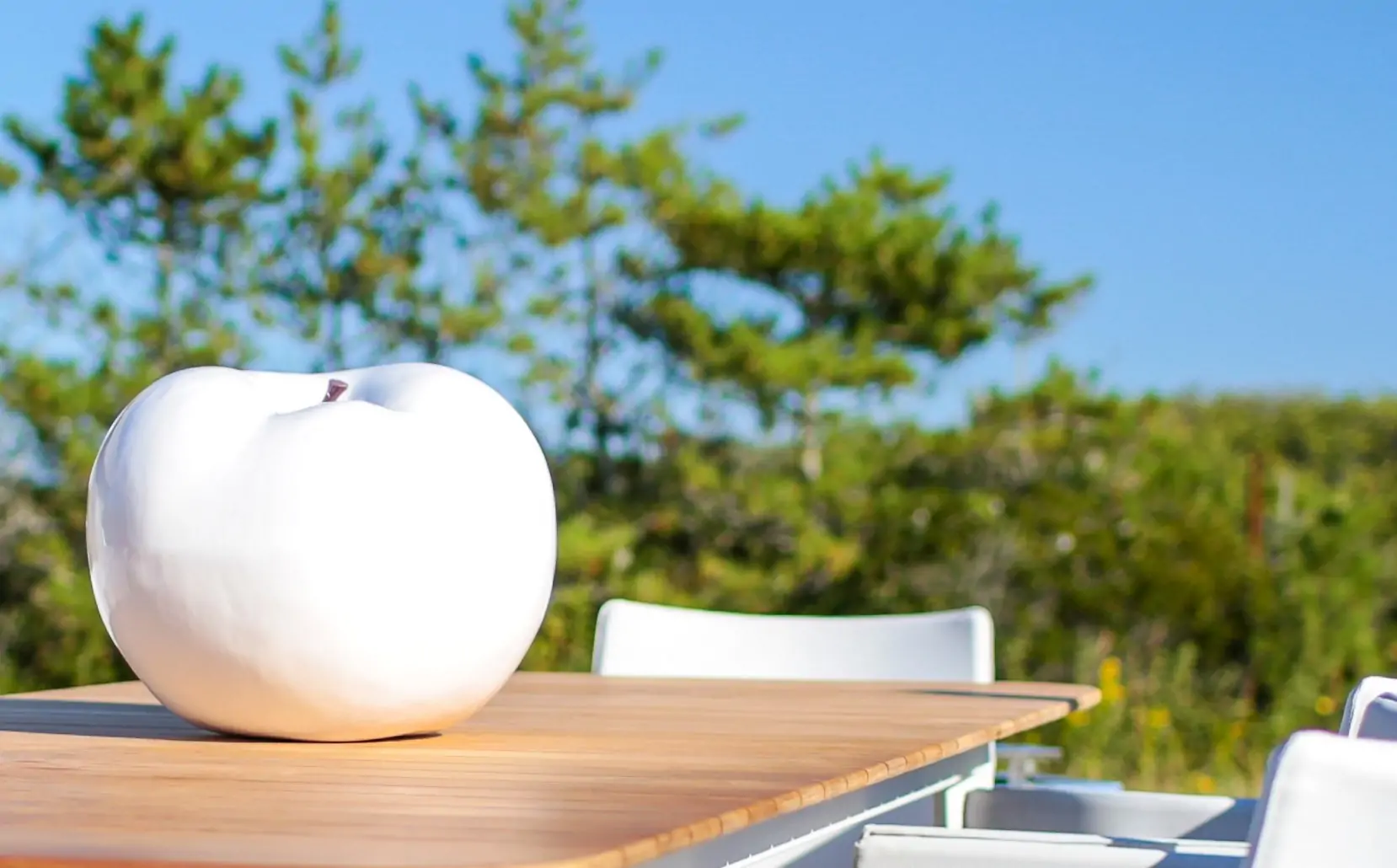 Giant white apple sculpture on an outdoor wooden table with trees in the background.